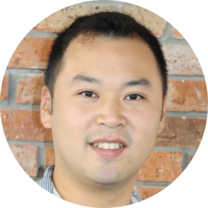 An asian man smiling in front of a brick wall.