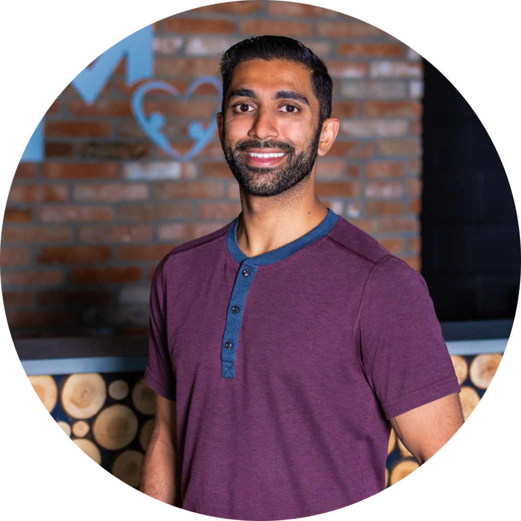 A member of our team, wearing a maroon shirt, stands smiling in front of a brick wall and wooden log background.