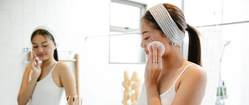 Woman in a white tank top, who recently joined a skincare-focused healthcare membership, applies product with a cotton pad in front of the bathroom mirror, wearing a headband.