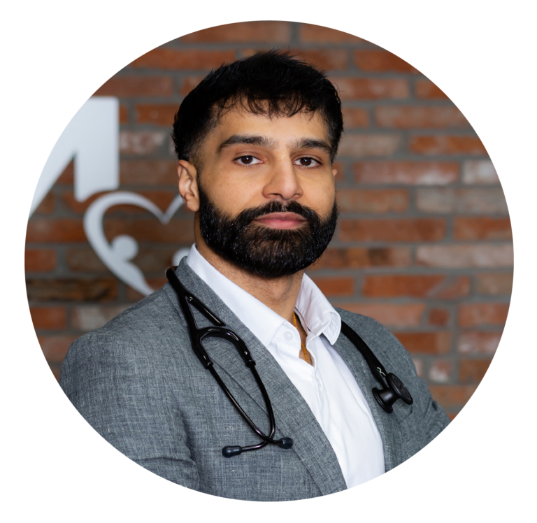 A professional portrait of a bearded male doctor from our team wearing a gray blazer and stethoscope, standing in front of a brick wall with a medical sign.