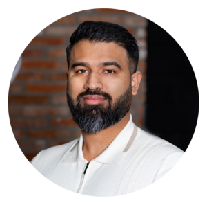 Our Team: A professional portrait of a man with a full beard, smiling, wearing a white shirt against a blurred brick wall background.