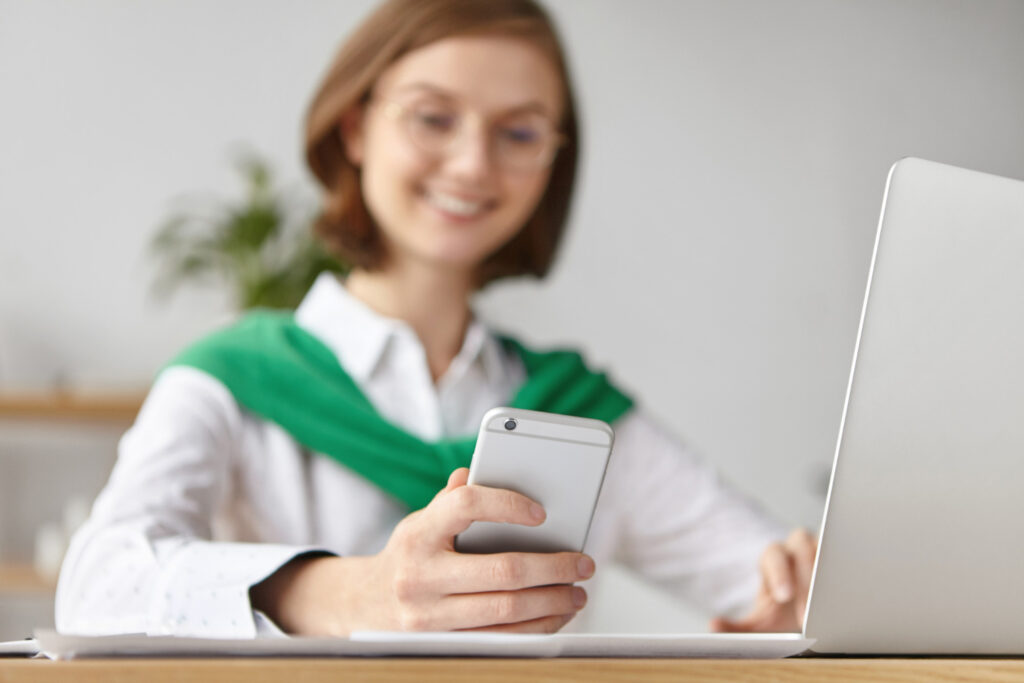 A person with glasses smiles while looking at their phone, possibly browsing the latest updates from Manhas Health Co. They are seated at a desk with an open laptop, wearing a white shirt and a green sweater draped over their shoulders.