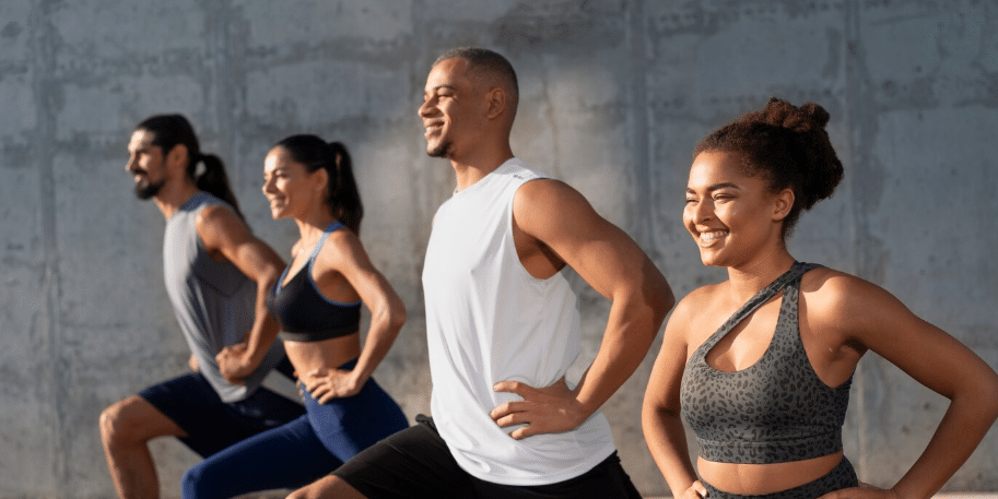 Four people in athletic wear exercise outdoors, standing in a row and smiling while performing lunges against a concrete wall—showcasing the confidence that comes from healthy living and Medical-Grade Skin Care.