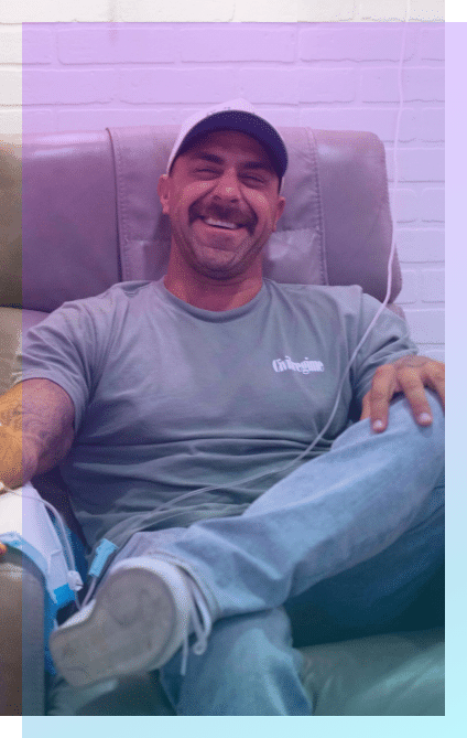 A man with a mustache, wearing a cap, gray T-shirt, and jeans, sits on a recliner chair smiling with his legs crossed, enjoying the comfort of Coquitlam IV Hydration Therapy.