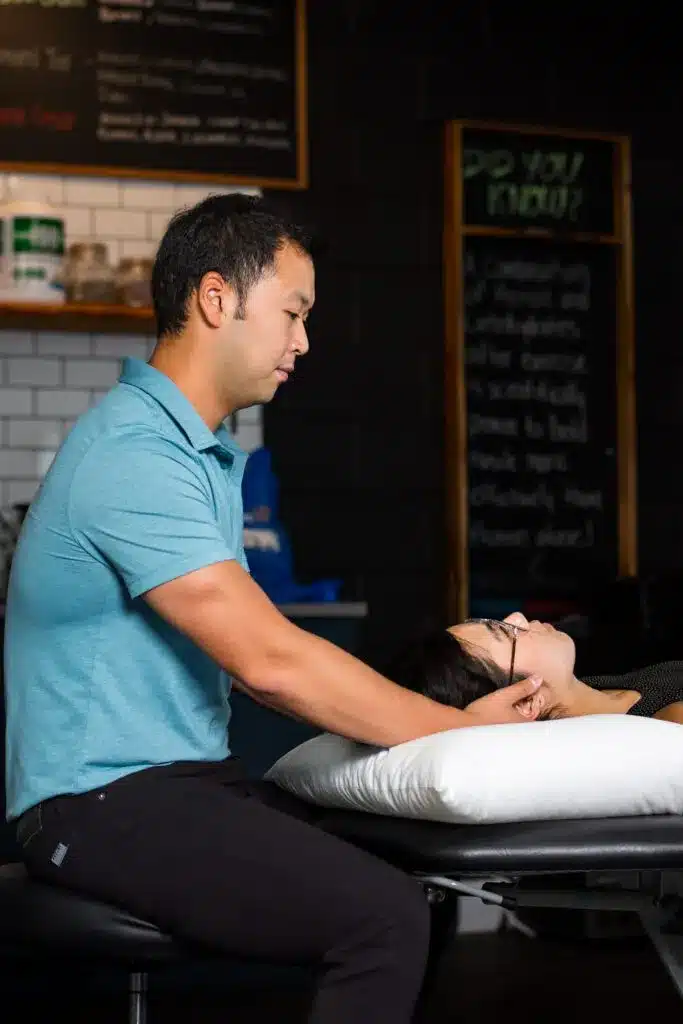 A man in a blue shirt from Manhas Health Co performs a neck adjustment on a person lying on a massage table in a clinic setting.