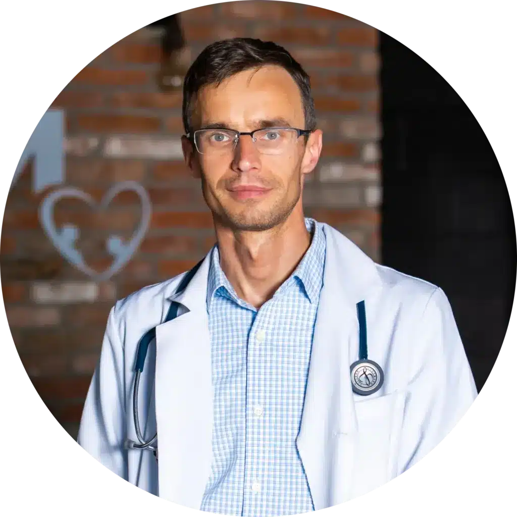 A member of Our Team, a man wearing glasses, a white lab coat, and a stethoscope stands indoors in front of a brick wall with a heart-shaped decoration.
