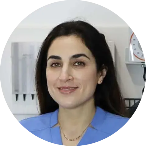 A woman with long dark hair wearing blue scrubs smiles while standing in a medical office, reflecting the professionalism and warmth of Our Team.