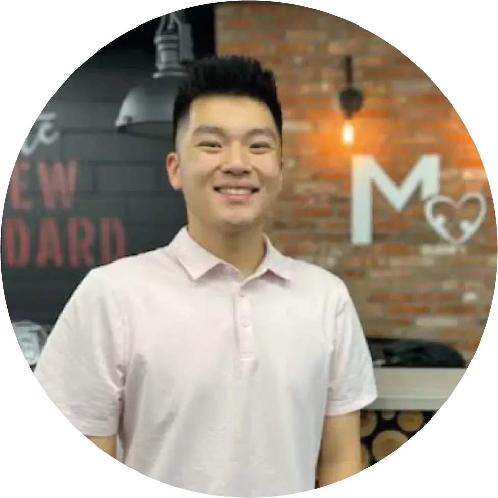 A member of Our Team stands indoors in front of a brick wall with a white "M" and heart symbol, wearing a light pink polo shirt.