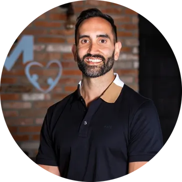 Man with short dark hair and beard smiling, wearing a black polo shirt, standing in front of a brick wall with a heart and arrow design in the background—proudly representing Our Team.